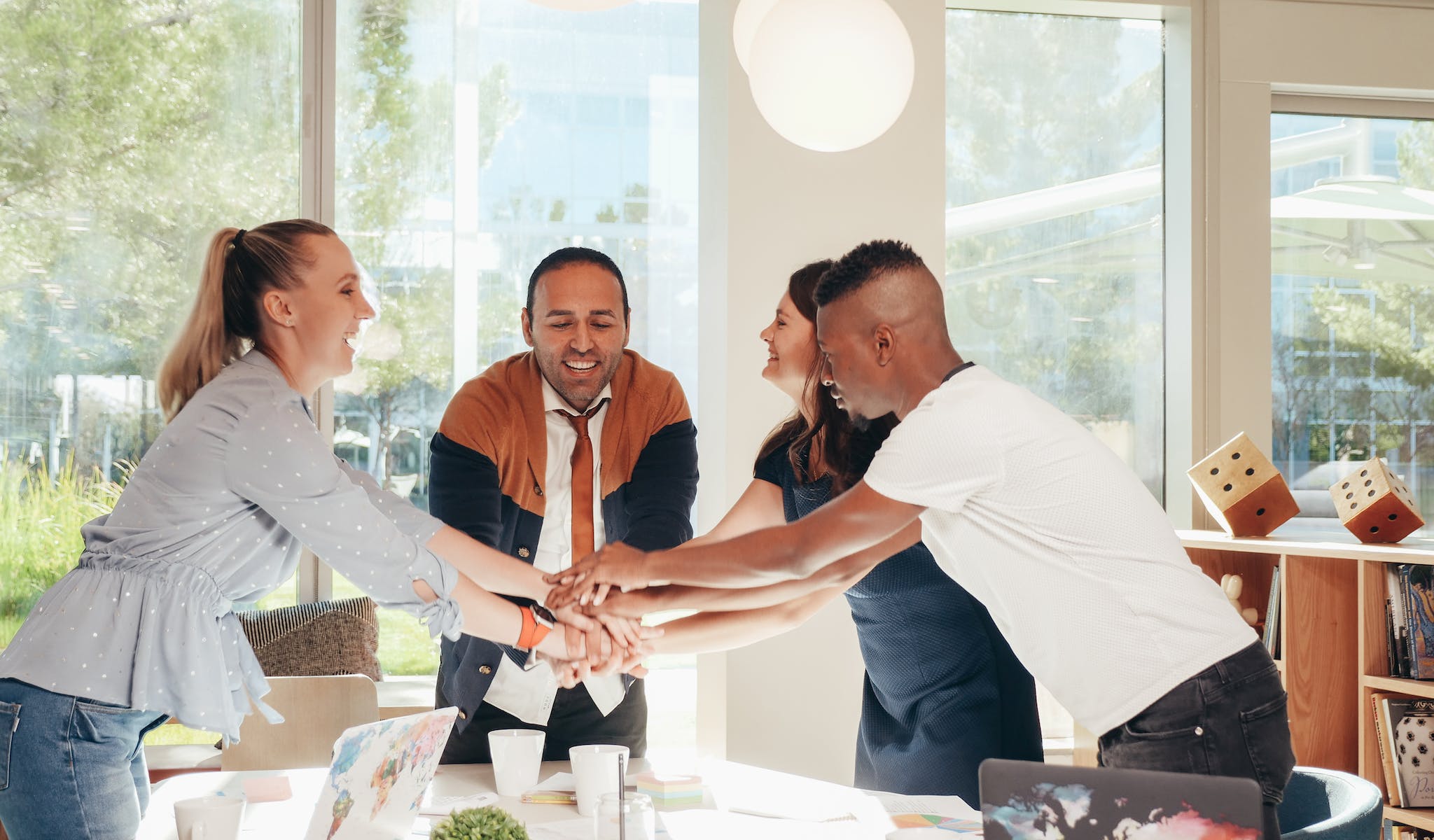 Content multiethnic colleagues joining hands above table with laptops in modern office on sunny day